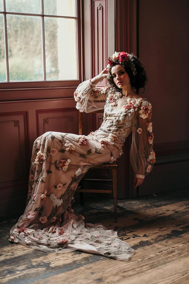 Young woman wearing long gown decorated with many applied flowers sitting by a panelled window. 