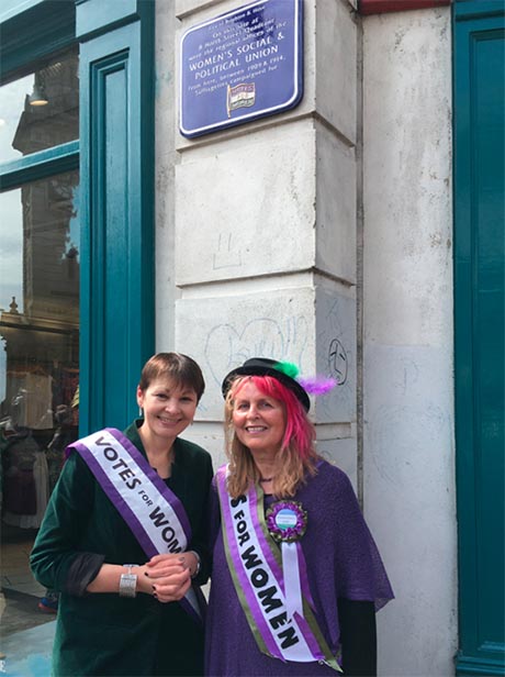 Brighton Pavilion MP Caroline Lucas and BHWHG Co-Secretary Maria Hogg, both wearing 'Votes for Women' sashes stand beneath a commemorative plaque to the WSPU