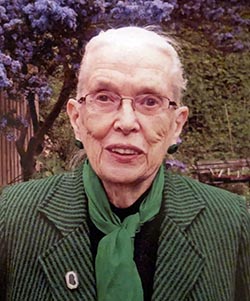 Outdoor photograph of an elderly woman wearing a green and black striped jacket, with a green scarf tied loosely around her neck.