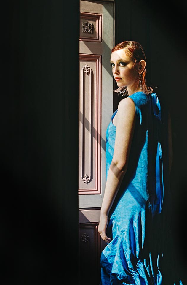 Photo of young woman standing by a window, she has red hair and is wearing a blue dress. 
