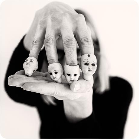 Black and white photograph showing a close up of a hand reaching towards the viewer, with the four fingers each inserted into downwards a head of a small doll