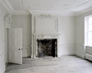 Photograph showing white-painted room with decorative cornice, doorway and fireplace