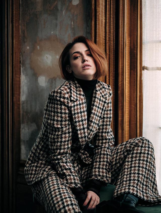 Photo of a young woman seated by a window, she is wearing a check trouser suit, in the background is a bare plaster wall.