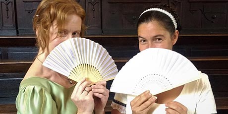 Photograph of two women in Regency costume, peering over the top of their fans