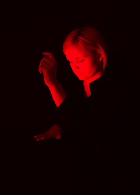 Photograph of a woman in black in a dark room, her hands and face illuminated by a red light