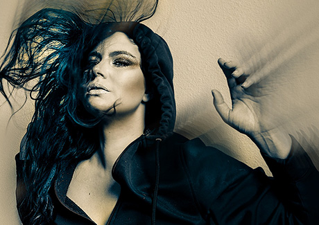 Head and shoulders photo of a woman with black hair and clothes, her hair is flying and her hand a blur of motion.