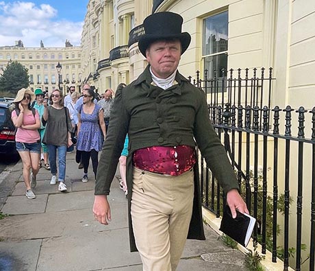 A man in Regency costume leading a tour group past a row of cream coloured town houses
