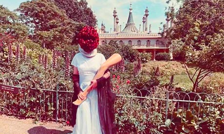 Photograph of a woman standing in the Pavilion gardens. She is wearing a white gown, carries a mannequin leg and a red floral mask obscures her face.