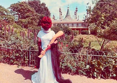 Photograph of a woman standing in the Pavilion gardens. She is wearing a white gown, carries a mannequin leg and a red floral mask obscures her face.
