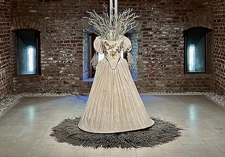 A full length 1665-style dress, stuffed and surrounded by bunches of lavender, displayed in a bare brick room and flanked by two slit windows.
