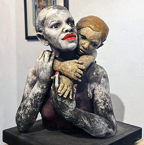 Photo of ceramic sculpture. It depicts the upper half of a woman set on a dark wooden plinth. A child clings to her back, its arms linked around her neck.