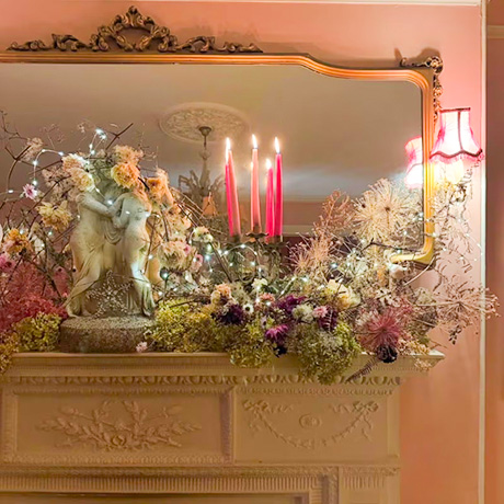 Photo of mantlepiece, decorated with flowers, cherubs, candles and a mirror behind.