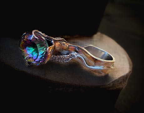 Photo of a piece of jewellery, a ring of twisting organic form set with a stobne that reflects blues, greens and purples